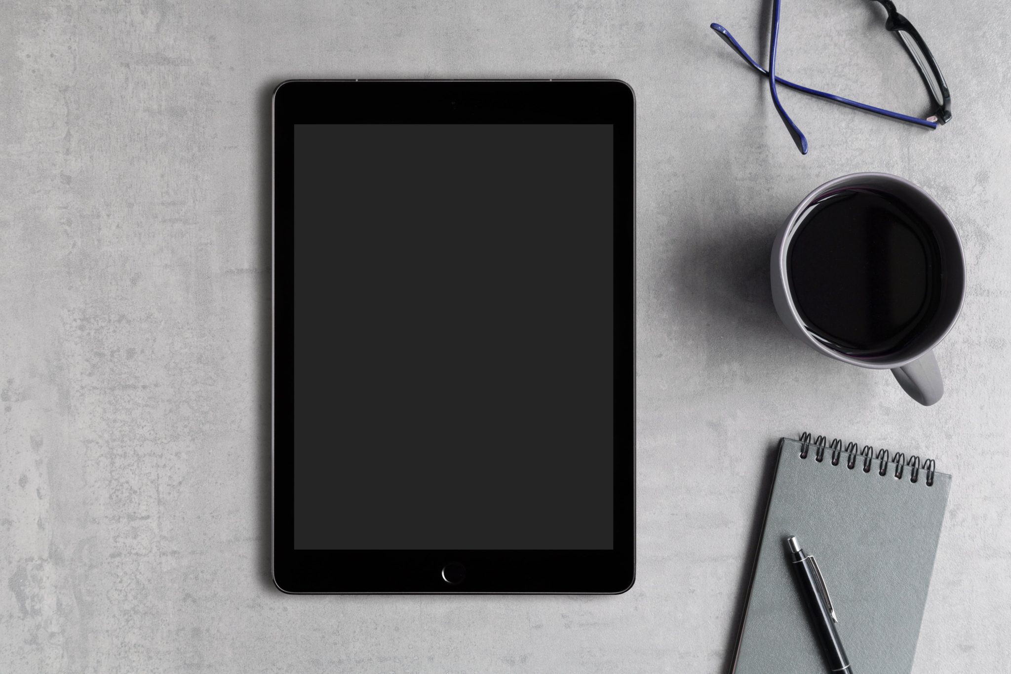 A black tablet, with a coffee cup, black glasses, a small notepad and a pen. All these office items are on a gray background.