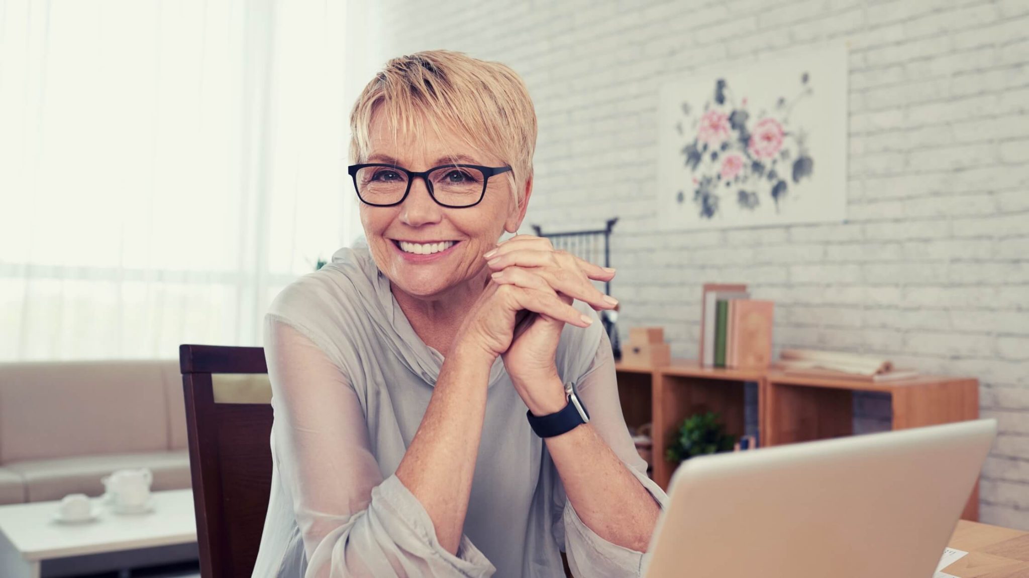 blonde woman with glasses, short hair, smiling, in front of her laptop, over 40 years old.