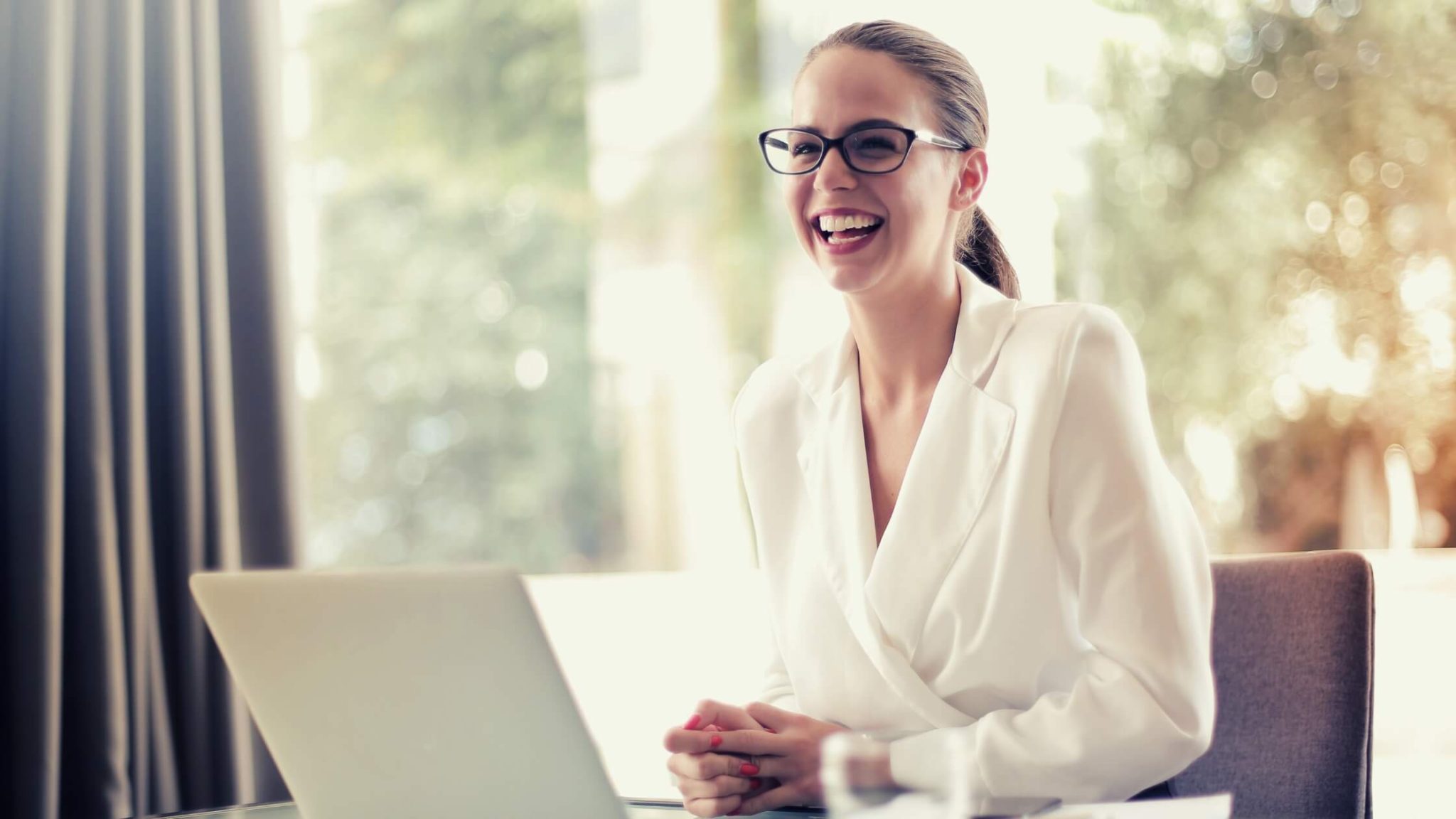 Well-dressed woman with glasses, smiling in front of her laptop.